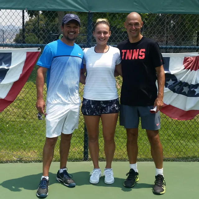 Ana Bogdan between Leon Bax and another coach on a sunlit court, bunting along the fence behind them.