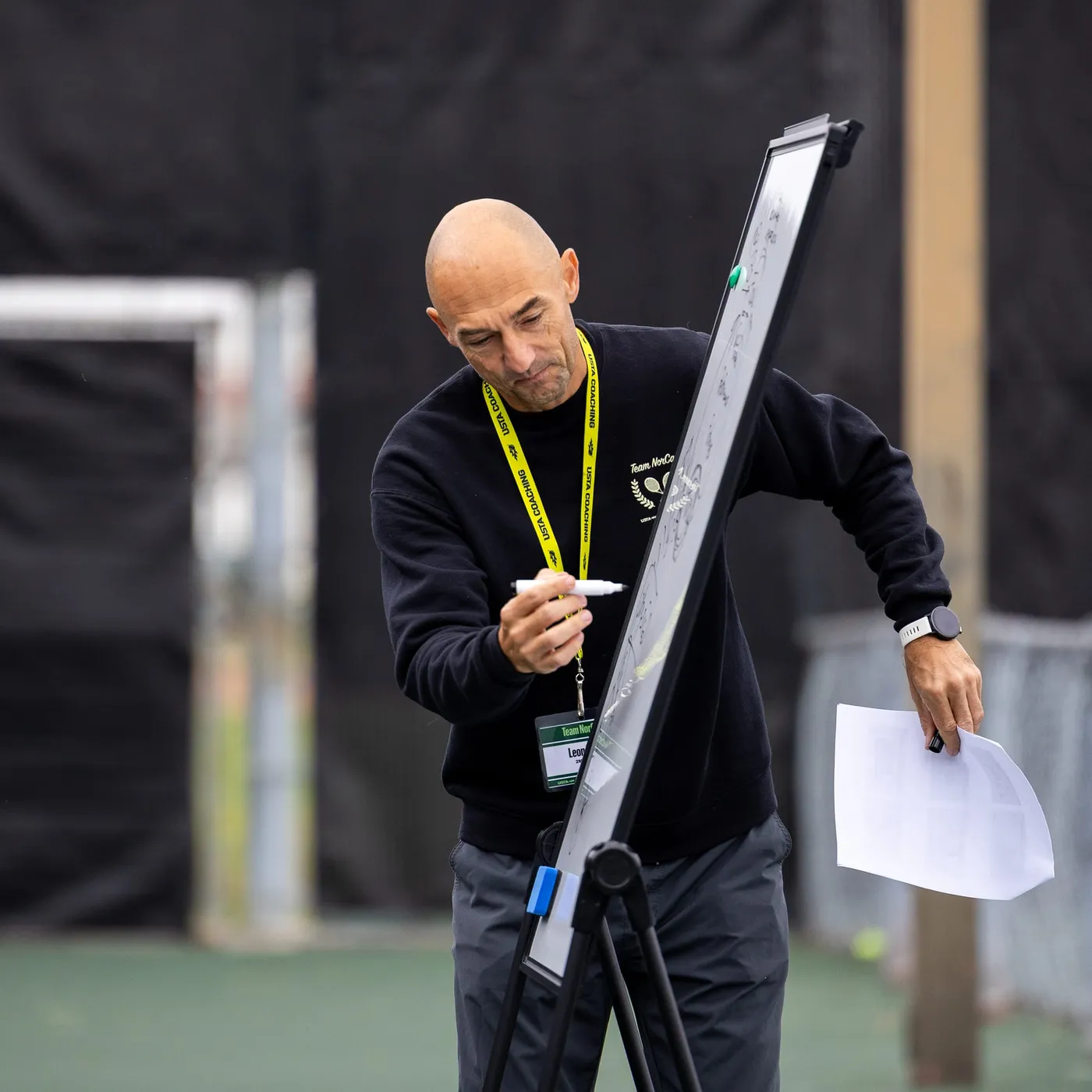 A coach writing on a flip chart beside a tennis court, a marker in one hand and a sheet of notes in the other.