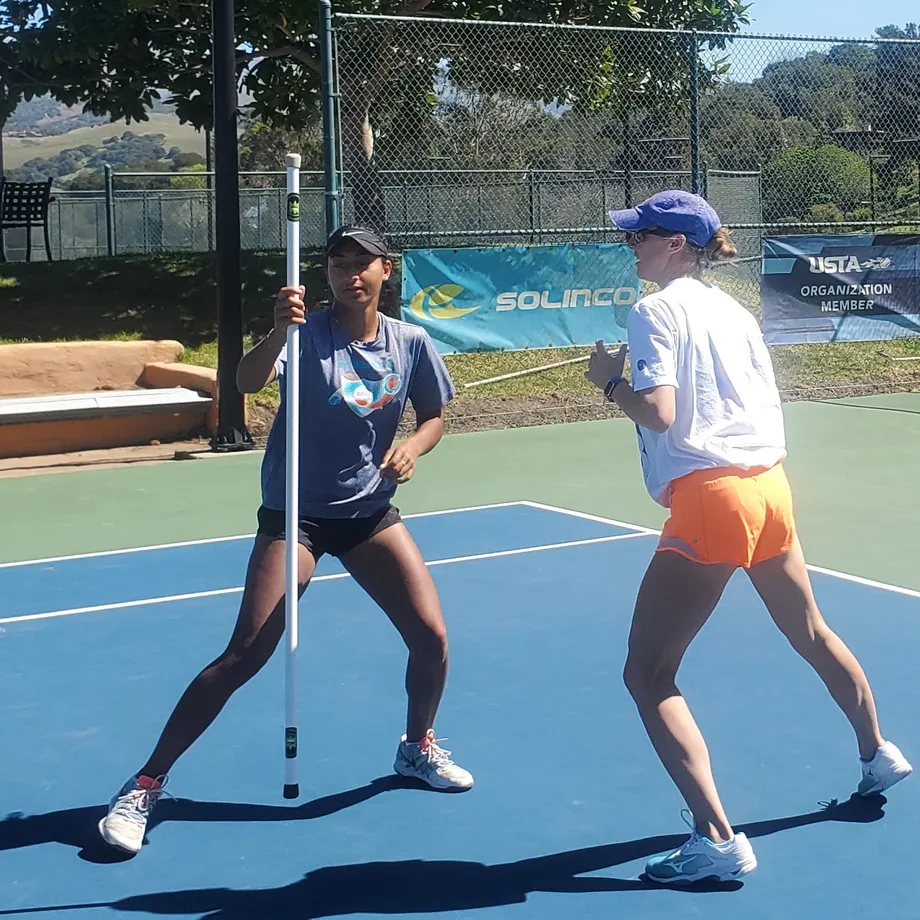 Two players on a hard court in low athletic stances either side of an upright stick, mid-drill.