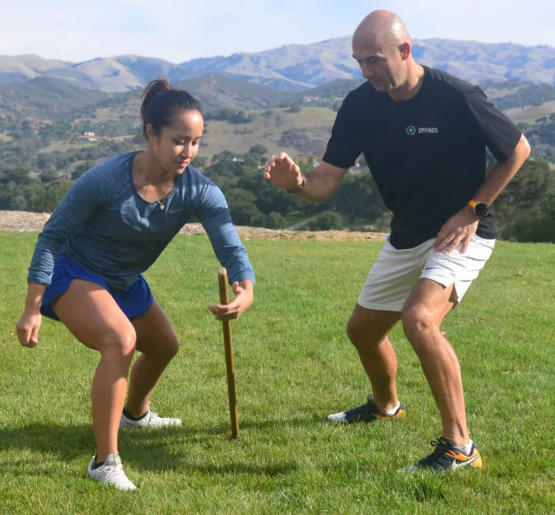 An athlete in a low athletic stance on grass, eyes down on an upright stick in front of her, with a coach alongside, hand raised beside the stick.