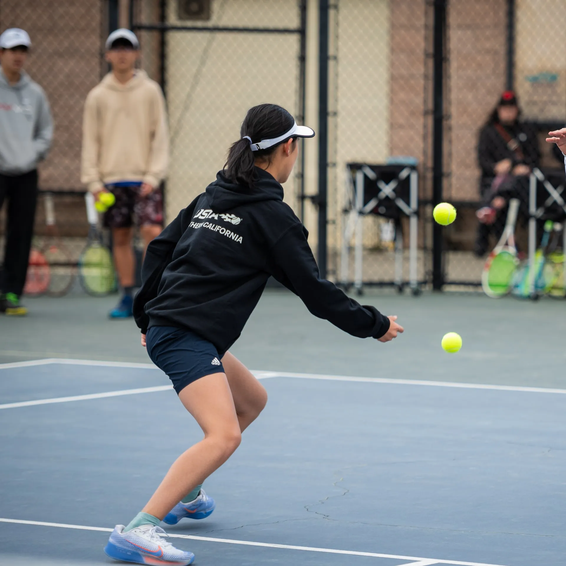 A junior player moving to a ball during a court drill, a coach feeding in front of her.