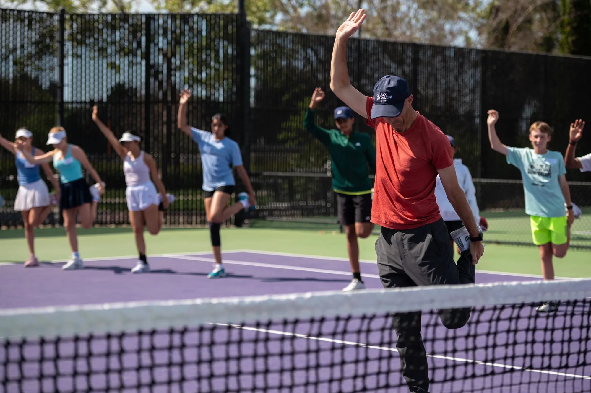 A coach leading a line of young players through a standing quad stretch on outdoor courts.
