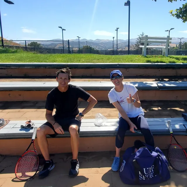 Kaitlyn Christian and Leo Prieto seated on courtside bleachers with their racquets, hills behind them.