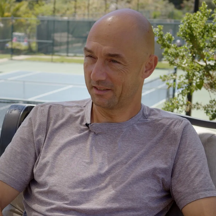 Leon Bax seated courtside in conversation, tennis courts behind him.