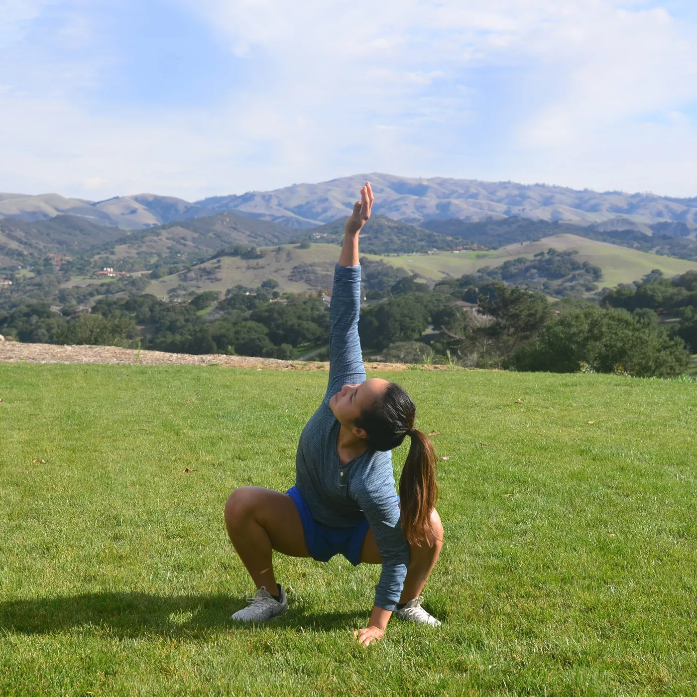A woman mid-stretch on open grass, one arm reaching up, rolling hills behind her.