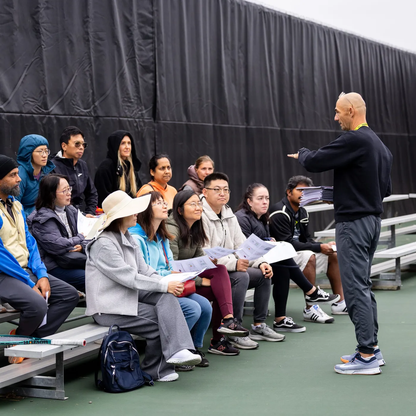 Parents sitting on courtside bleachers with printed handouts, listening to a session being led in front of them.