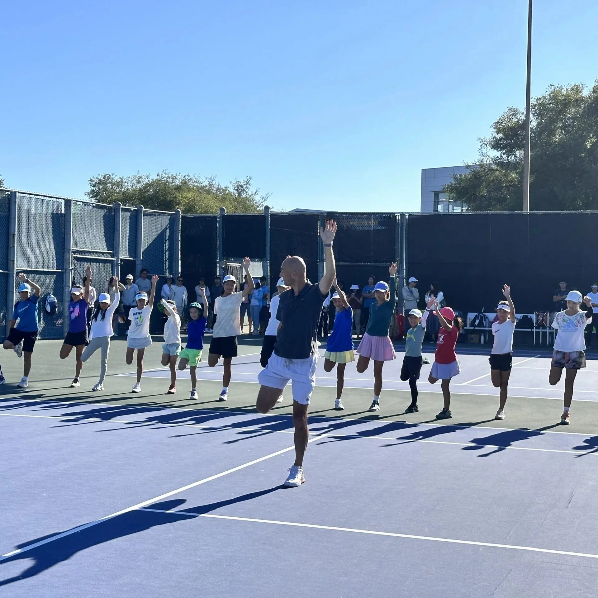 Leon Bax leading a large group of children in a standing balance stretch on outdoor courts, with parents watching behind.